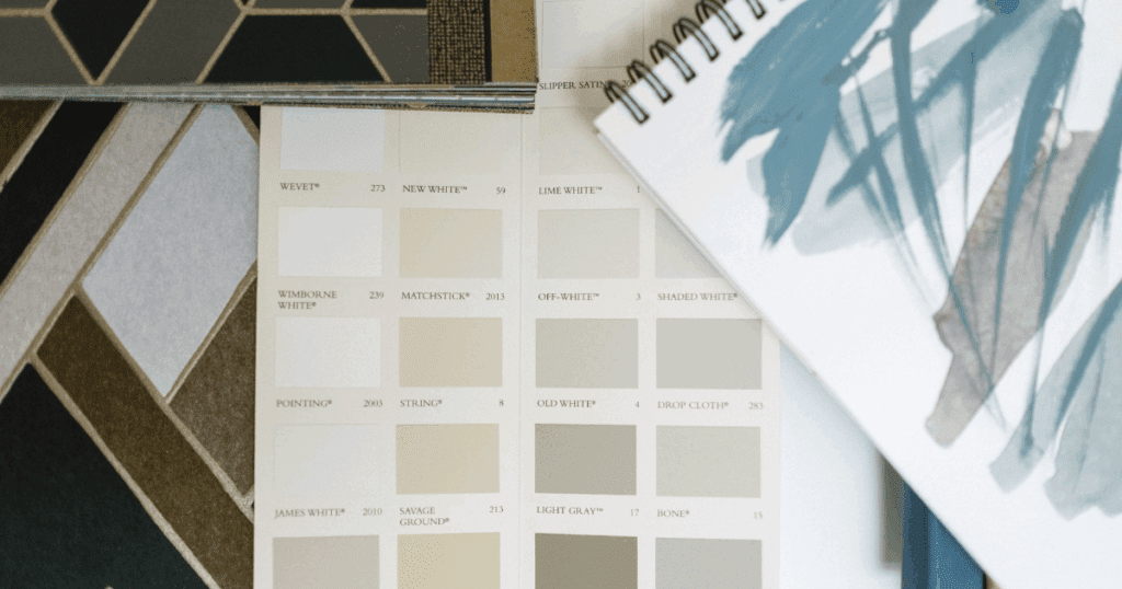 A detailed paint swatch book featuring off-whites, creams, and greys, paired with fabric samples and a sketchbook with watercolor designs, laid out on a design board.