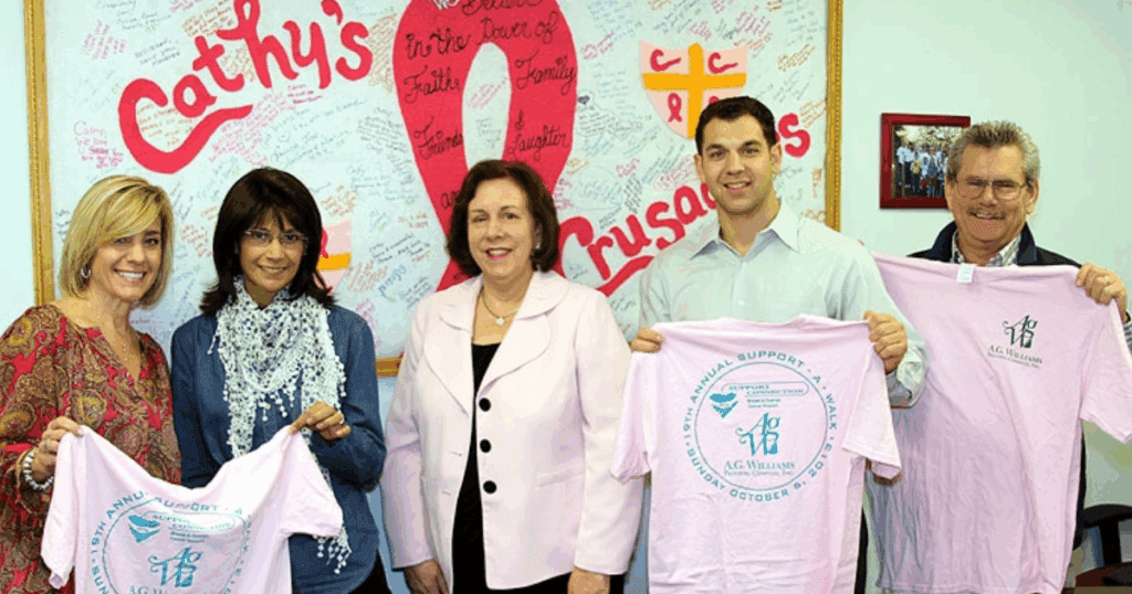 Five people standing together smiling and holding pink A.G. Williams Painting Company t-shirts in front of a breast cancer awareness tribute wall.