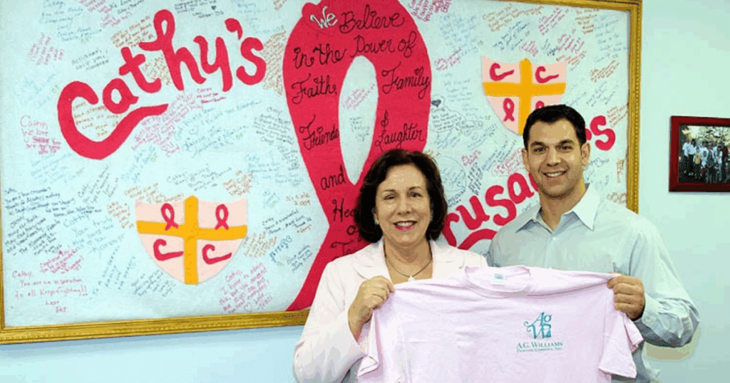 Two people smiling and holding a pink A.G. Williams Painting Company t-shirt in front of a breast cancer awareness banner that reads "Cathy's Crusaders."