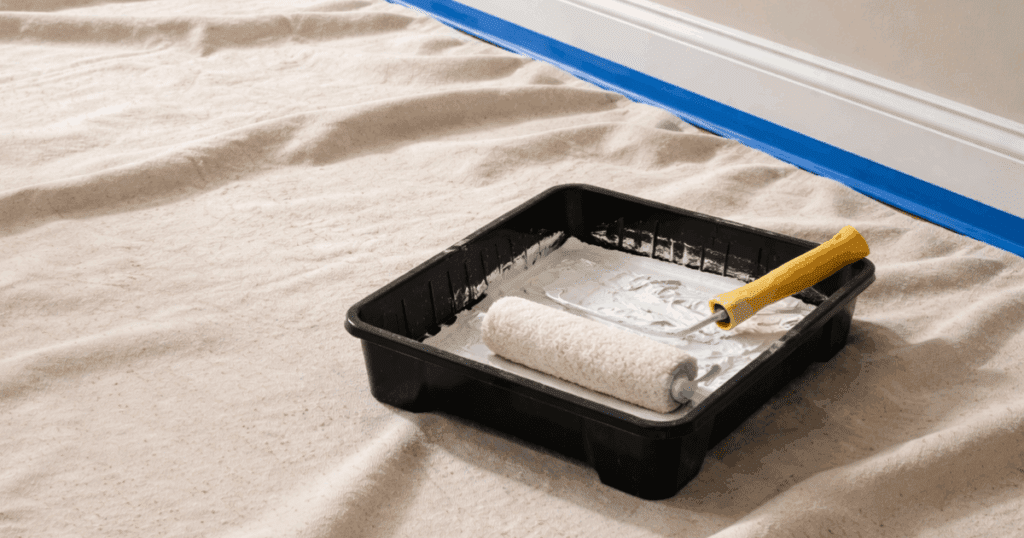 Paint roller resting in a tray of white paint on a drop cloth-covered floor with painter’s tape along the baseboard.