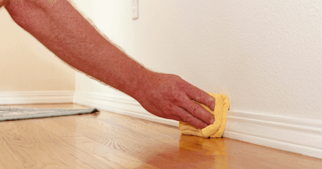 Close-up of a hand using a yellow cloth to clean a white baseboard along a freshly painted wall and hardwood floor.