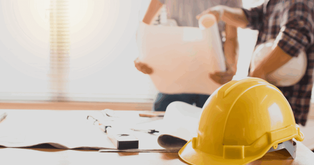 Close-up of a yellow hard hat on a desk with construction plans and two contractors discussing blueprints in the background for a blog about the importance of working with an insured painting company.