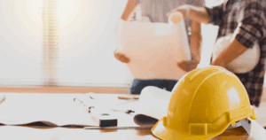 Close-up of a yellow hard hat on a desk with construction plans and two contractors discussing blueprints in the background for a blog about the importance of working with an insured painting company.