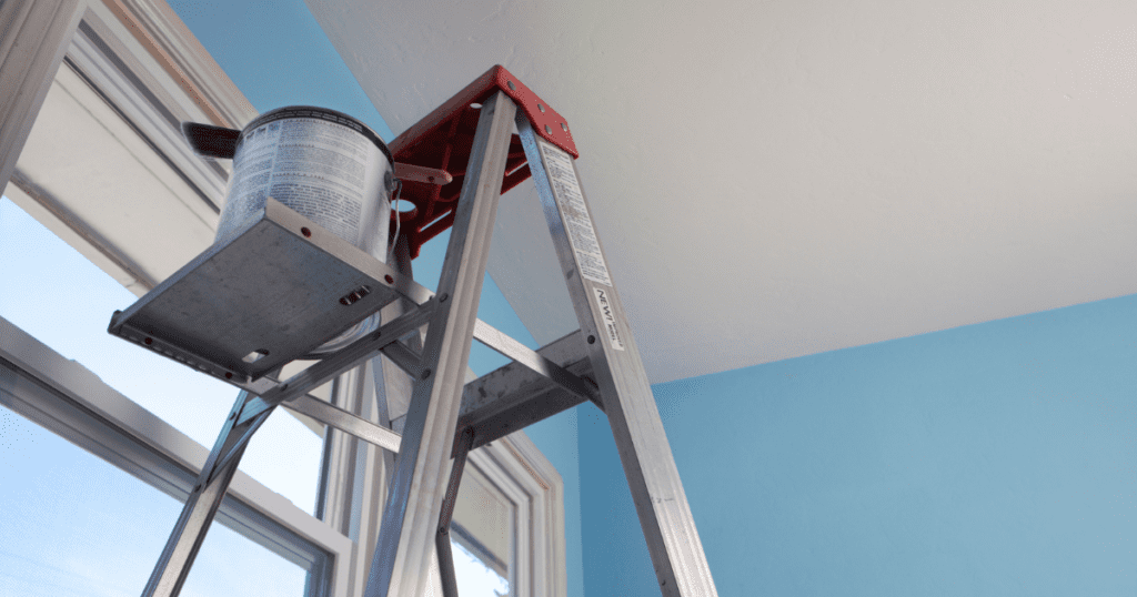 Step ladder with ceiling paint and brush set up in a blue-walled room with natural light.