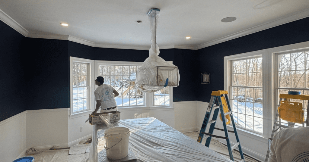 Professional painters prepping a navy and white room for ceiling painting with ladders and protective coverings.