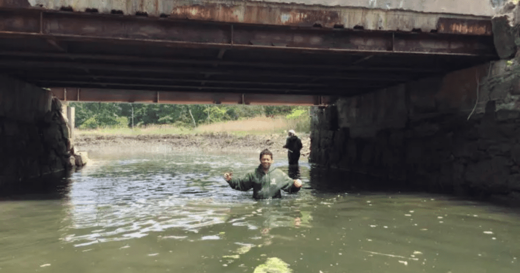 Painting crew standing in shallow water beneath a bridge inspecting and preparing rusted steel beams for restoration.