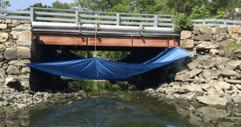 Bridge over water with a suspended containment tarp system installed beneath to capture debris during painting preparation.