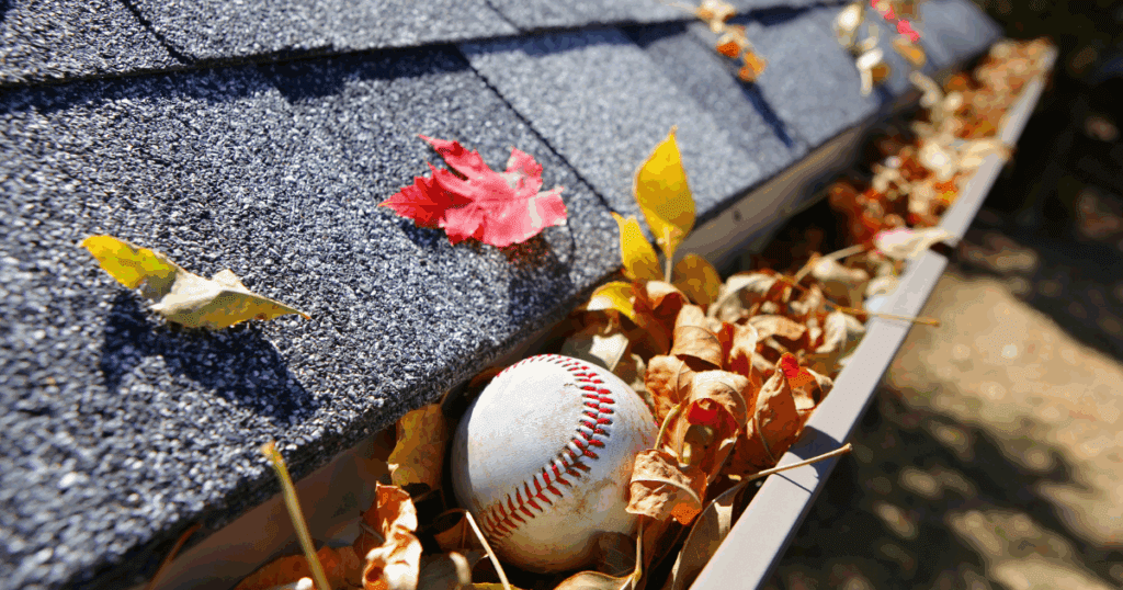 Gutter filled with autumn leaves and a baseball, emphasizing the need for clean gutters to avoid clogs and water damage.