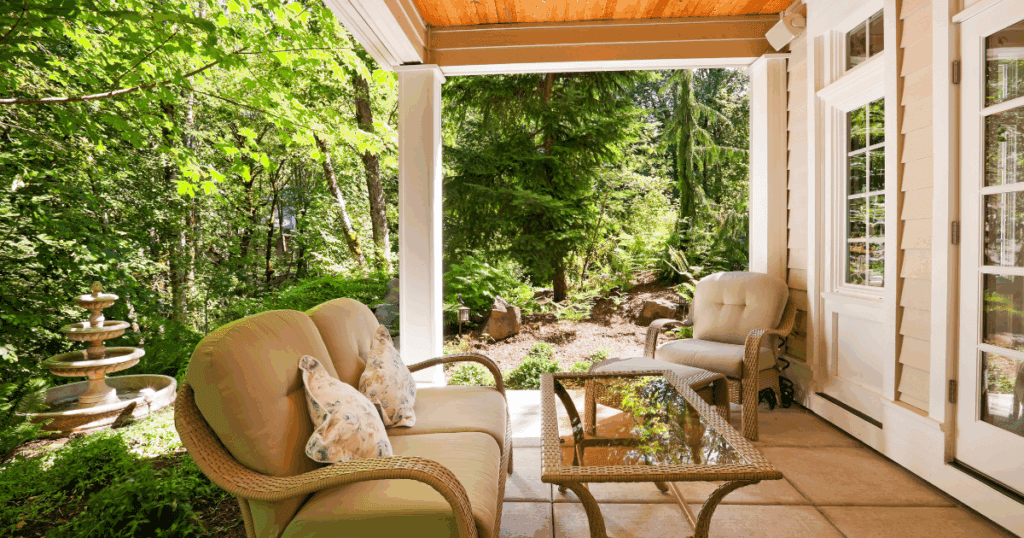 A peaceful covered porch with light-colored trim and siding, featuring comfortable wicker seating and surrounded by lush woodland.