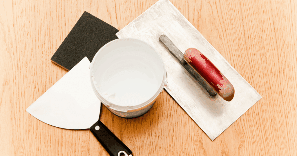 Drywall repair tools including joint compound, a putty knife, sanding sponge, and a finishing trowel on a wooden surface.