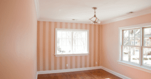 A small room with a large window, featuring peach-colored walls and a striped accent wall in the same shade. The room is empty, with natural light flooding through the windows, highlighting the wooden floor.