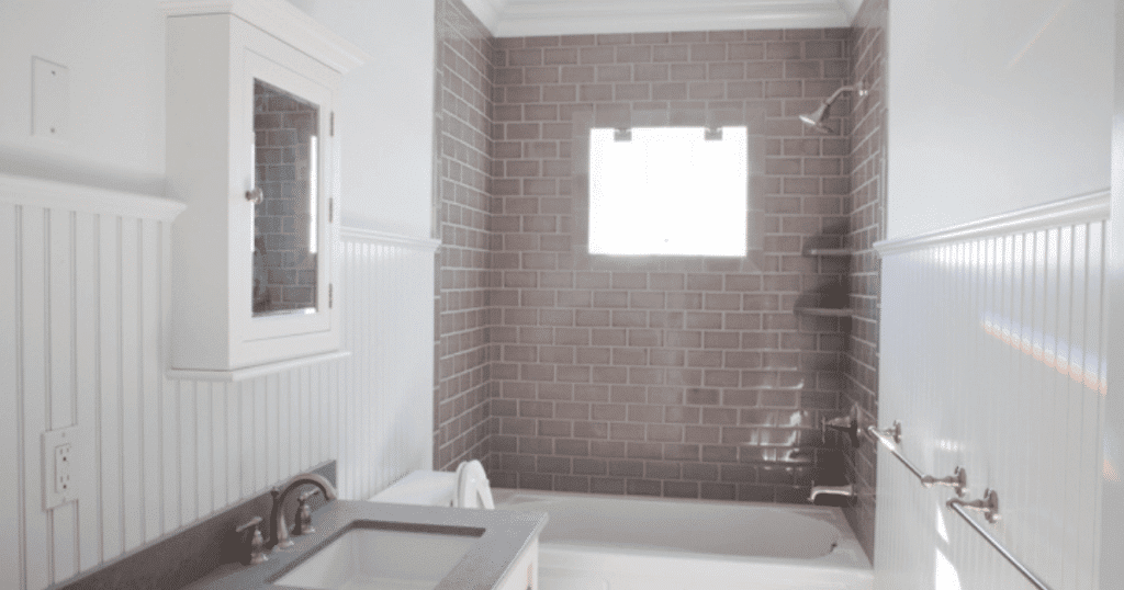 A bright, small bathroom with a gray subway tile shower wall and a white wainscoting panel on the lower half of the walls. The room includes a white vanity with a gray countertop and a white medicine cabinet with a mirrored door.