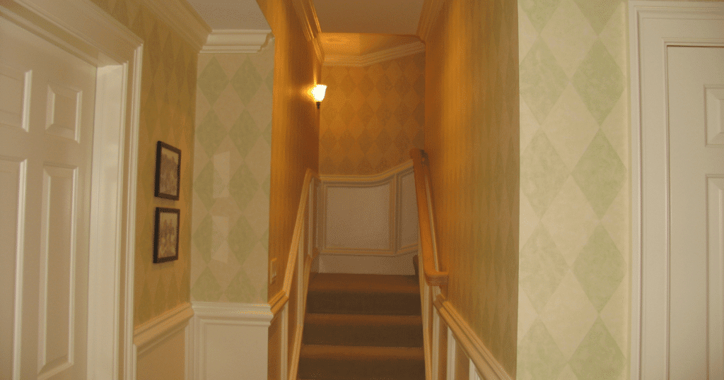 A narrow stairwell with light green and beige diamond-patterned wallpaper, featuring white wainscoting panels and a wooden handrail. Soft lighting from a wall sconce enhances the warm tones.