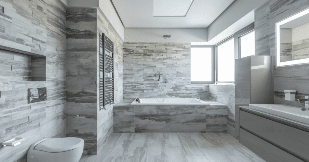 A contemporary bathroom with gray stone tile walls, a built-in bathtub, painted gray walls, and large windows allowing natural light.