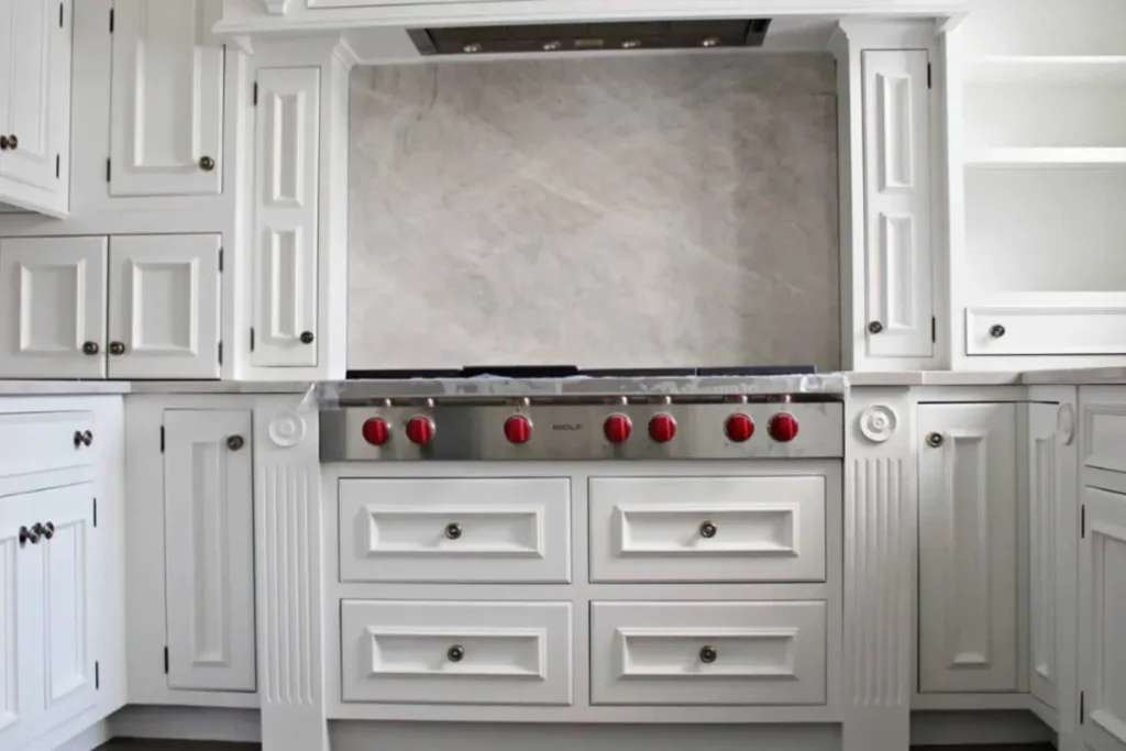 A bright and elegant kitchen with freshly painted white cabinetry featuring detailed paneling, polished chrome hardware, and a modern stove with vibrant red knobs. A testament to the transformation that occurs when you hire a professional kitchen cabinet painter.