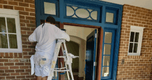 Painter applying fresh blue and brown paint to an elegant wooden front door with sidelights and transom.