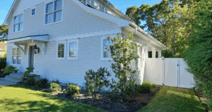 Light gray shingle-style home with clean white trim and lush landscaping under a clear sky.