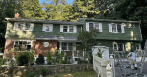 Painters working on a large two-story home with green shingles and white shutters.