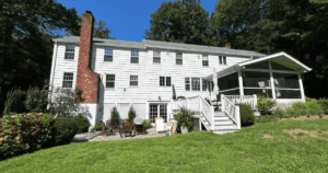 Large white shingle-style home with brick chimney and screened porch, viewed from the backyard.