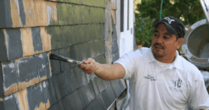 A painter applies fresh paint to weathered cedar shingles on a home exterior.