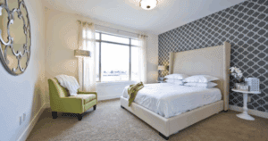 Bright bedroom with patterned wallpaper accent wall, beige upholstered bed, and green accent chair.