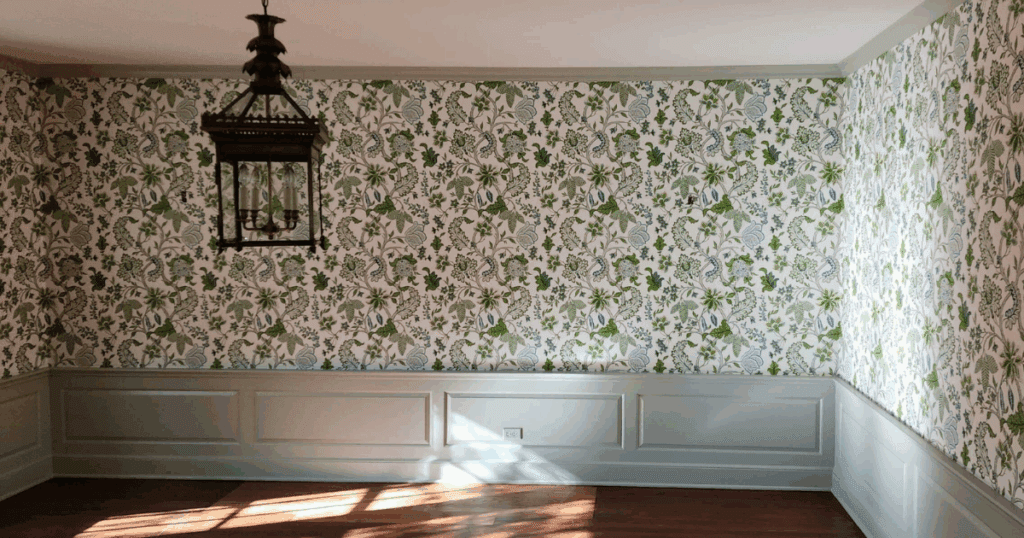 Sunlit dining room with green and blue floral wallpaper above soft gray wainscoting.