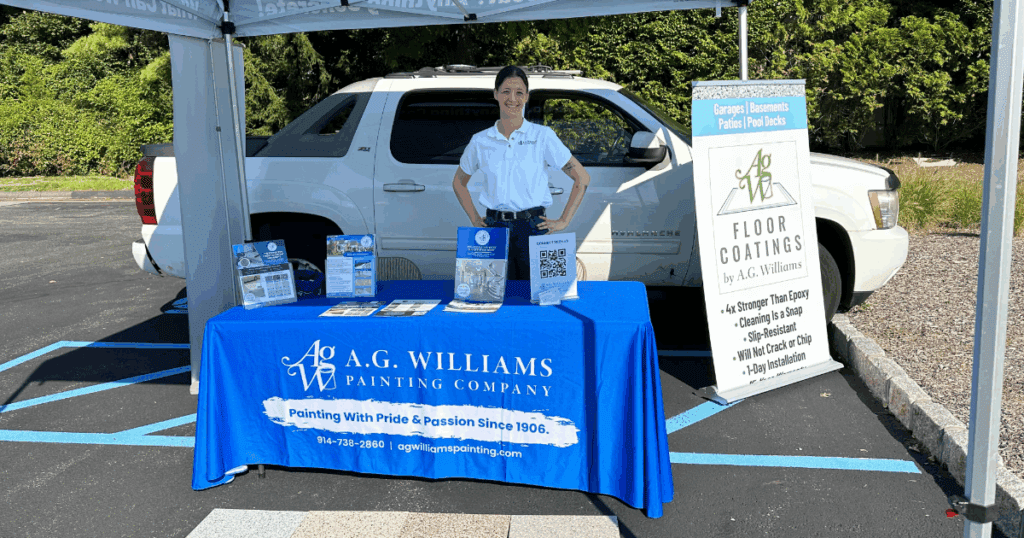 A representative from A.G. Williams Painting Company stands behind a branded blue table at an outdoor event, showcasing company materials and a display promoting floor coatings. A white SUV and promotional signage are visible in the background.