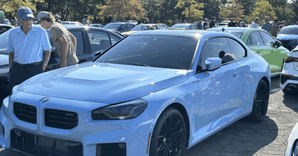 A sleek light blue BMW M2 is parked at a car show, surrounded by attendees and other vehicles under a sunny sky.