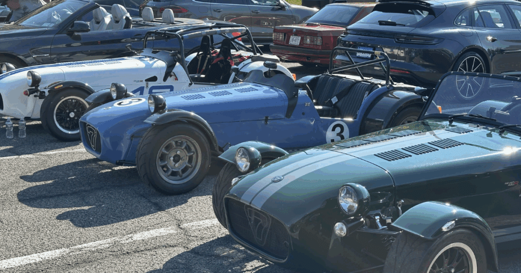 A lineup of vintage-inspired roadsters, including Caterham and Lotus 7-style cars in green, blue, and white, are parked side by side at a car show.
