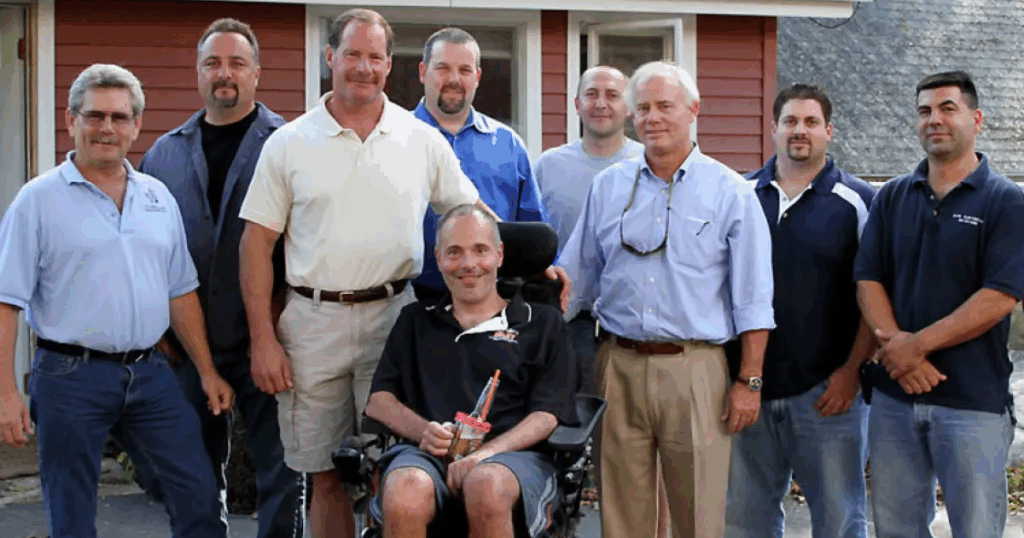Group photo of A.G. Williams Painting Company team members and community partners standing with Sergeant Roger Petrone, seated at the center, outside his restored Connecticut farmhouse during the Home in Need project.