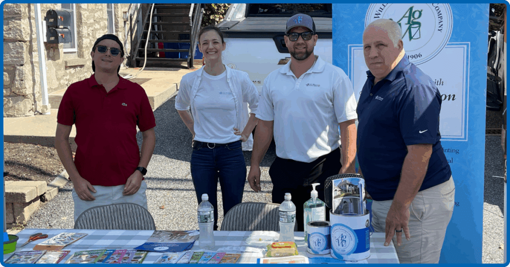 A.G. Williams Painting Company team members smile in front of their booth at Mt. Pleasant Day, standing beside product displays and a company banner.