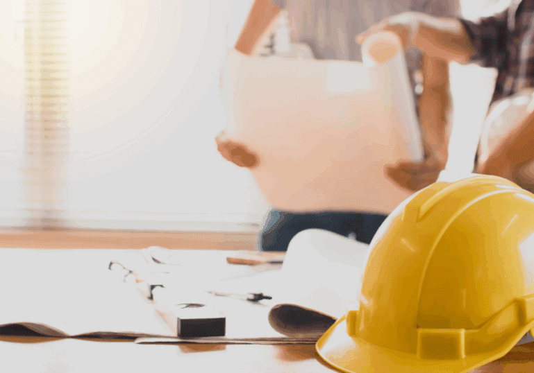 Close-up of a yellow hard hat on a desk with construction plans and two contractors discussing blueprints in the background for a blog about the importance of working with an insured painting company.
